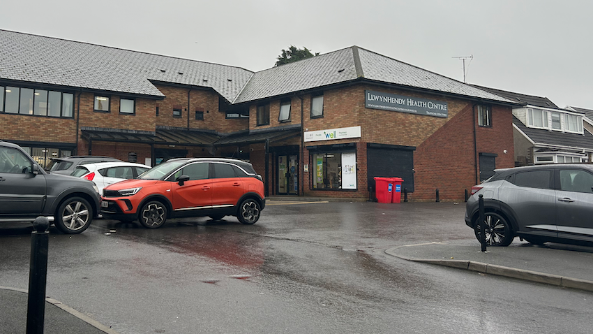 Llywynhendy health centre is a brick building with a sign. There is a car park in the foreground with several cars parked. 