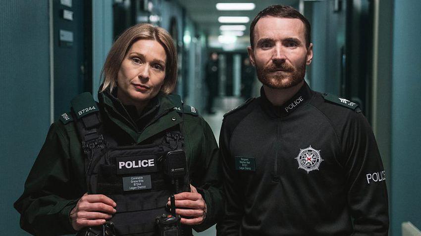 Sian Brooke and Martin McCann stand as their characters Grace Ellis and Stevie Neill in the corridor of a police station looking at the camera. They are wearing police uniforms, with Sian Brooke on the left wearing a large black vest with her name and station code. 