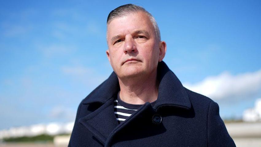 Craig Jones on a beach wearing a navy blue coat and blue striped top, with grey hair