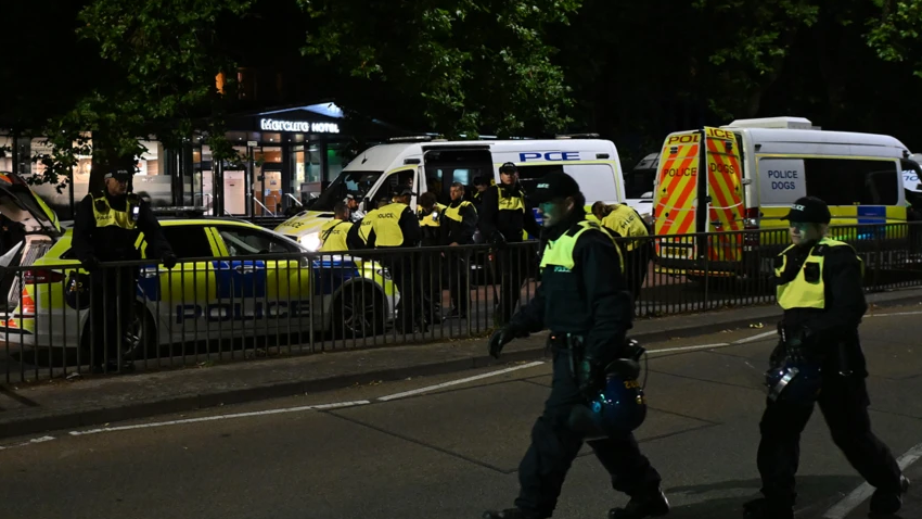 Police walking down the road in Bristol in riot gear. In the back of the image two police vans and a police car are parked in the road.