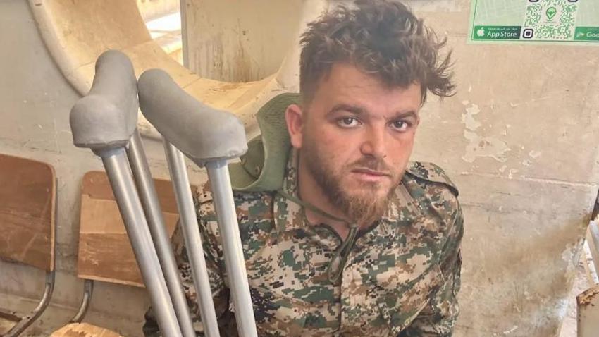 A young man dressed in a camouflage combat uniform holding crutches stares at the camera. His face is dirty and his eyes look tired.