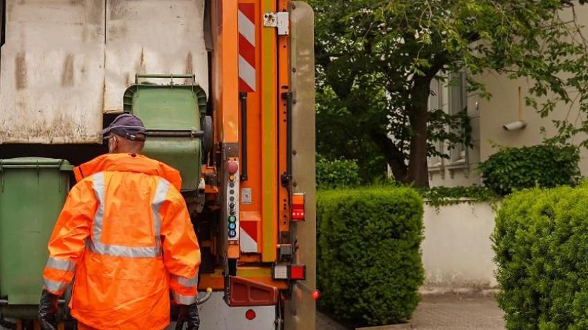 A refuse collector wearing an orange jacket, standing in front of a bin lorry with his back to the camera