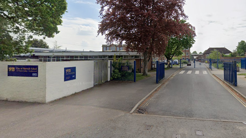 The outside of City of Norwich School. The building is set back from the road and has blue gates. On a white painted building is a blue sign which says "City of Norwich School" in yellow writing.