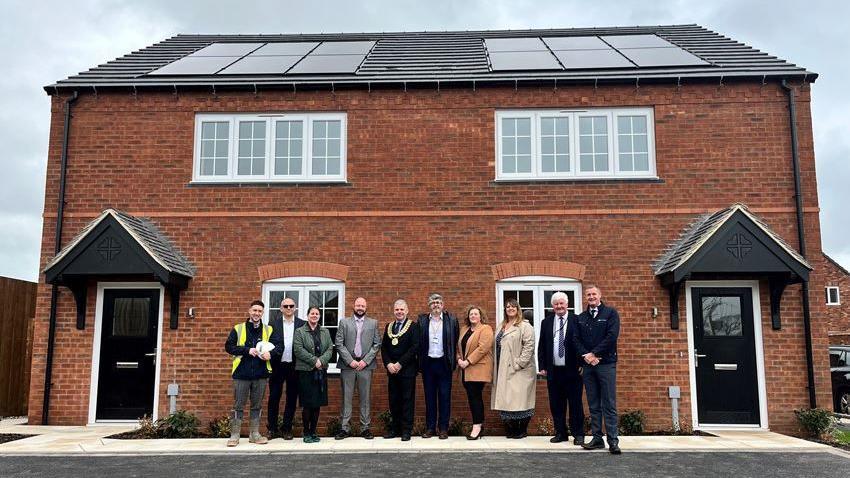 A total of 10 people including councillors and construction bosses stand in front of two three-bed houses. They have solar panels on the roof.