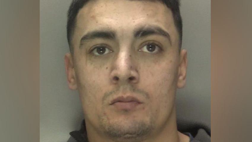 A police mugshot shows a man with short brown hair and light stubble. He is wearing a dark grey top.