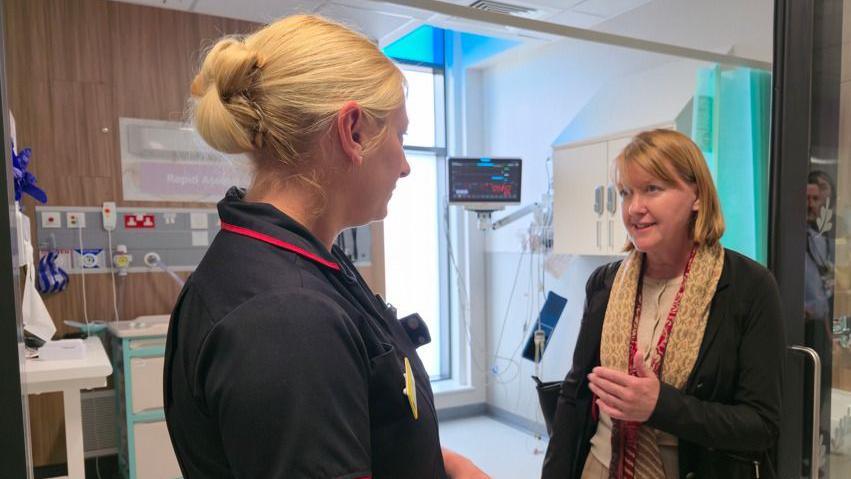 Sarah Gibson wearing a beige cardigan underneath a black jacket, with a cream patterned scarf over the top. She is standing in a dentistry ward, speaking to a woman with blonde hair who is wearing black scrubs.