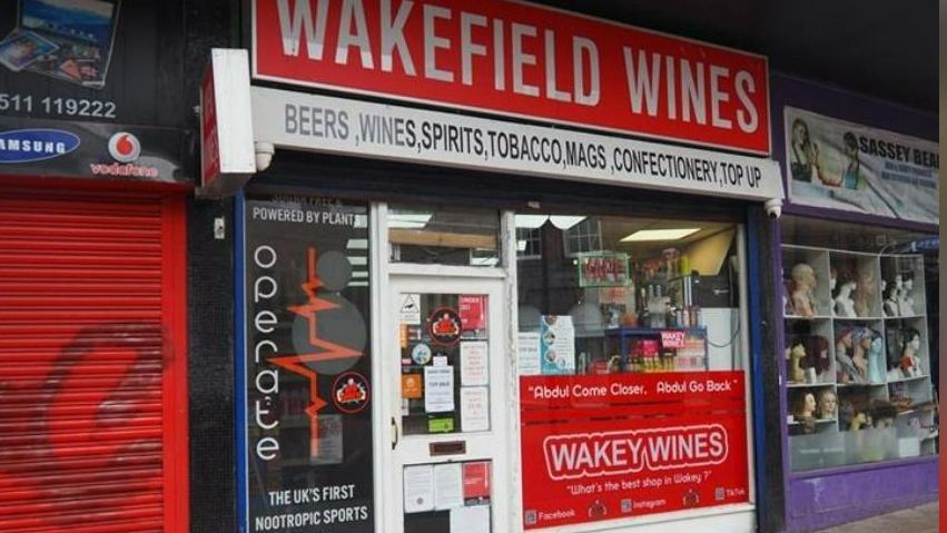 A red sign has white writing saying 'Wakefield Wines', with a white sign beneath it saying 'beers, wines, spirits, tobacco, mags, confectionery , top up'. Another sign in the window says 'Wakey Wines' and includes the catchphrase 'Abdul come closer, Abdul go back'.