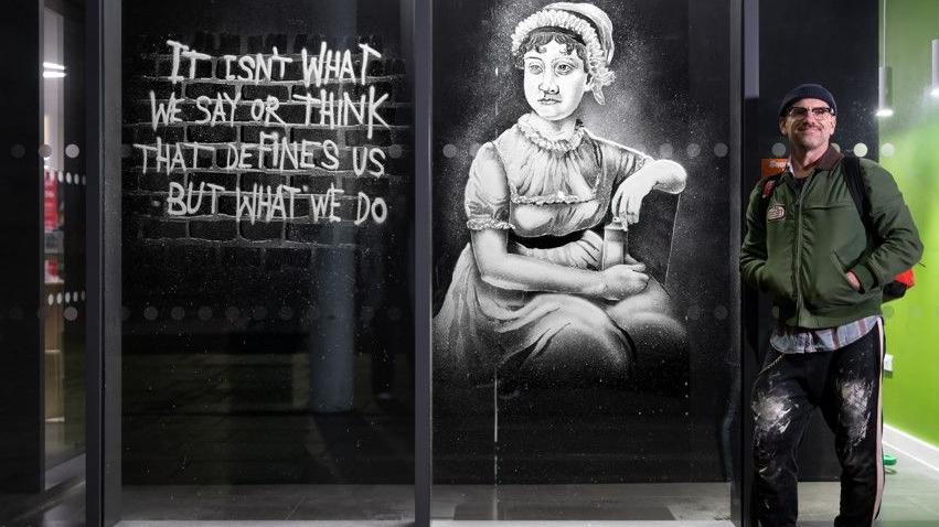 Man with green jacket with his hands in his pockets standing outside by a window decorated with an image of Jane Austen sprayed with fake snow.