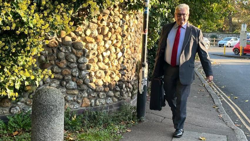 A full-length shot of Simon Spence walking along a pavement next to a road and a flint wall. He is carrying a case and is wearing a dark suit, blue shirt and a red tie. He has a full head of grey hair and is wearing glasses.