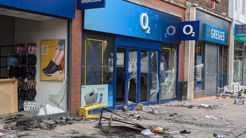 O2 frontage with smashed windows and doors. In front of the building there is rubbish and shards of glass strewn all over the floor. On the right is a Greggs store which has similar scenes but the shutter is down. On the left is a shoe shop with the window completely smashed and on the floor