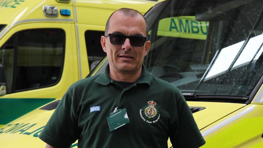 Stuart Little wearing a dark green polo top, with the Community First Responders' logo, and black sunglasses. He is standing in front of two ambulances.