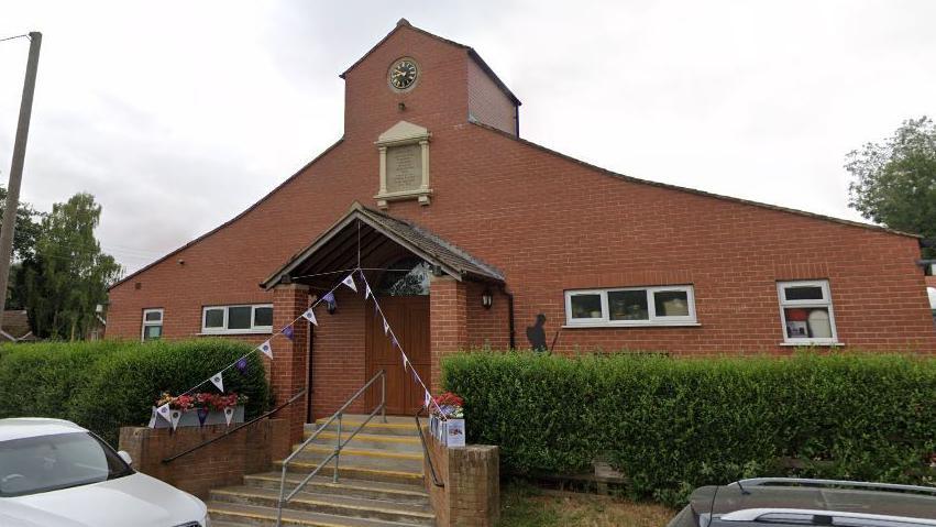 The exterior of Goxhill village hall. It has concrete steps leading up to the entrance, outlined with yellow paint and with a metal handrail in the middle. It has two lines of flags hanging from above the middle of the entrance. The building has small windows and large green bushes in front. Cars are parked on the road in front of the building. 