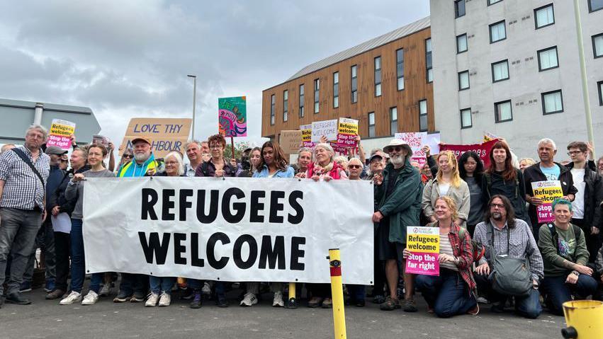 A group of people stand outside a large white and wood-panelled building. They hold a large sign reading "refugees welcome" as well as smaller signs with the same message. Another sign says "love thy neighbour".