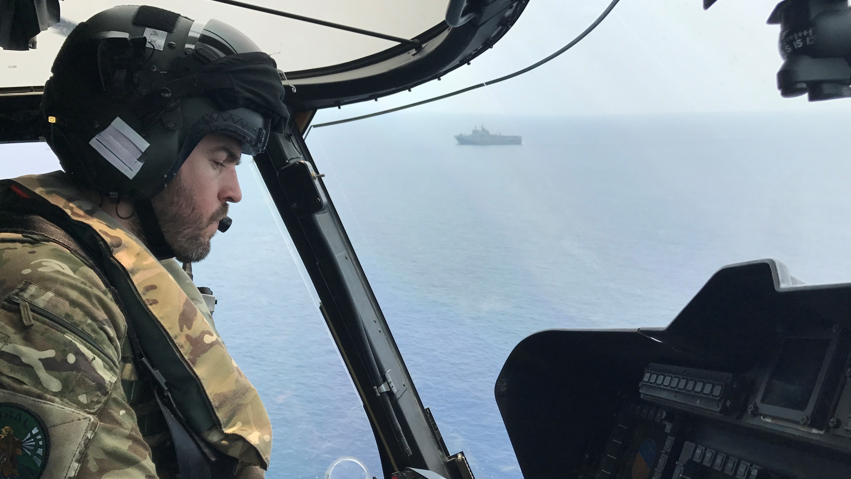 Steve Irwin in a helicopter above the sea wearing camouflage uniform and a helmet, with a military-type ship in the far background