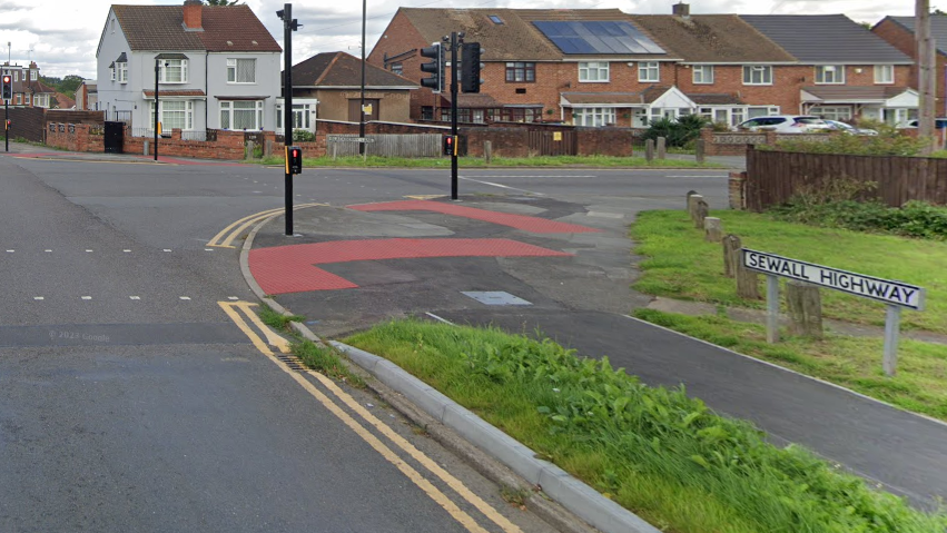 A road junction with the sign Sewall Highway in the foreground. Several houses can be seen beyond the junction with solar panels. The corner of the junction has pedestrian crossings across two of the roads