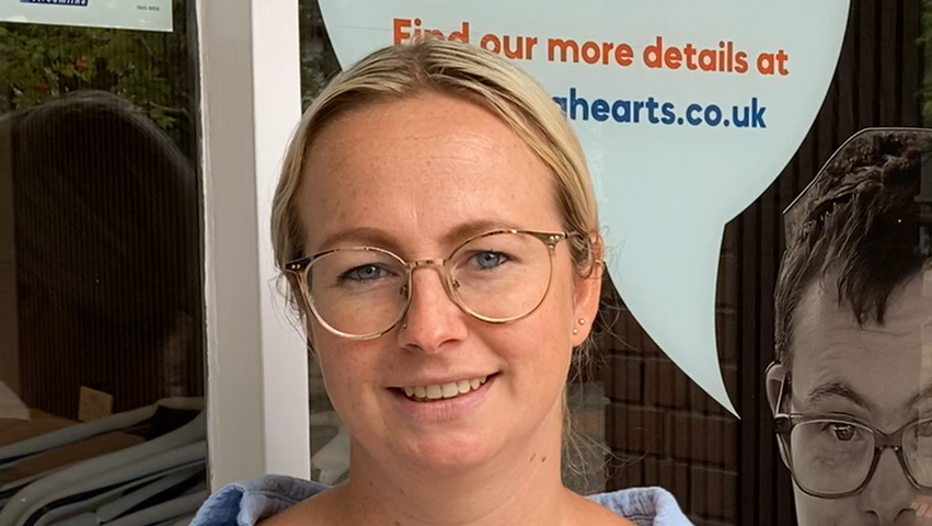 Emma Pulford smiles at the camera outside the cafe. She has blonde hair that has been tied back behind her head. She wears gold framed glasses and a light blue shirt. 