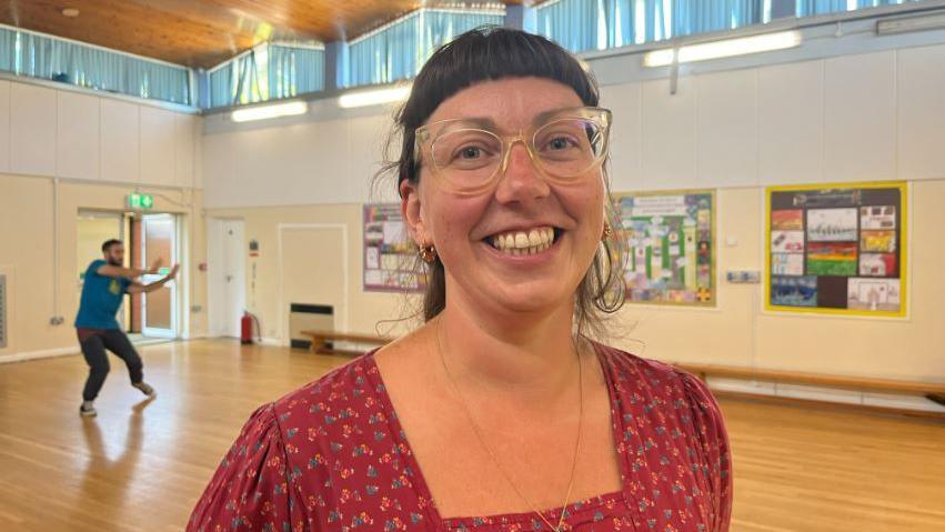  Robyn Cabaret smiling and looking into the camera in a school hall. She is wearing a floral pink dress and glasses.