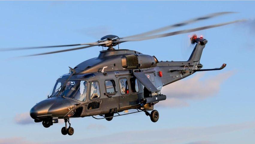 A photograph of an AW149 flying. There is a red tail light on and the wheels are down.