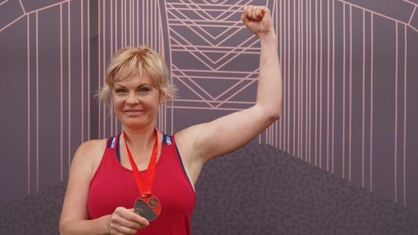 Marharyta Marchenko has blonde hair tied in a ponytail. She is wearing a red running vest and holding a medal in her right hand while flexing her muscles in a show of strength with her left arm. She is smiling towards the camera.