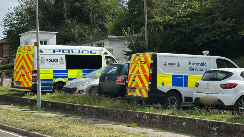 Two police vans - including a forensics services van - in a car park. Other cars are also in the image.