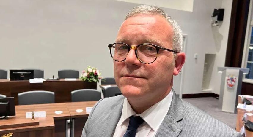 Will Russell looking side-on at the camera in a council chamber. He has short grey hair and leopard-print round glasses, and is wearing a white shirt, navy tie and a grey jacket.