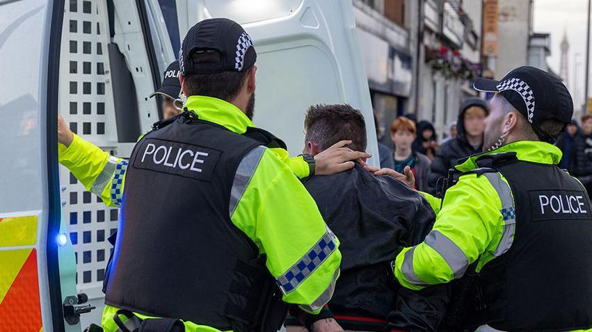 Man is arrested by three police officers and led into the back of a police van