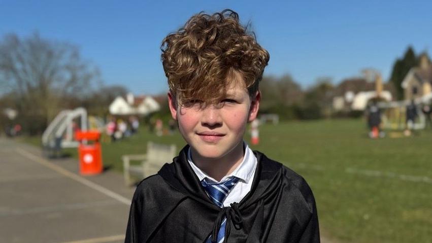 A schoolboy looking straight at the camera. He is wearing a white shirt, blue tie and black cloak and is standing in front of a playing field.