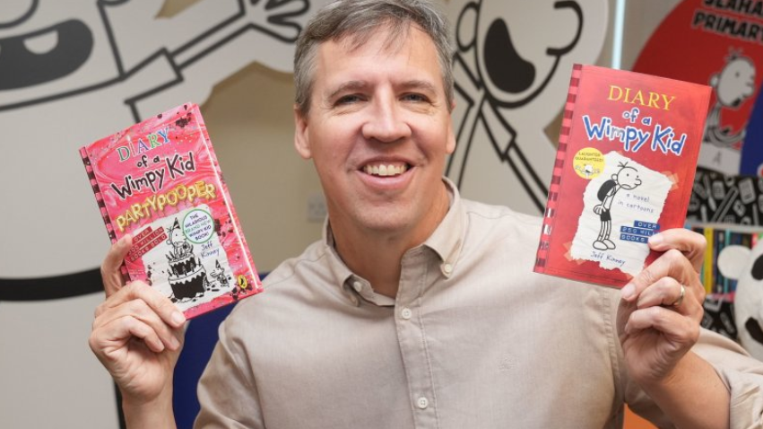 Kinney is smiling and holding two copies his Wimpy Kid books he is wearing a beige shirt.