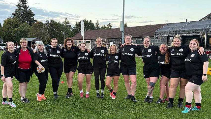 The Galts line up for a team photo. They are standing on a rugby pitch, all wearing a black kit, and they have their arms around each other. The clubhouse can be seen behind them.