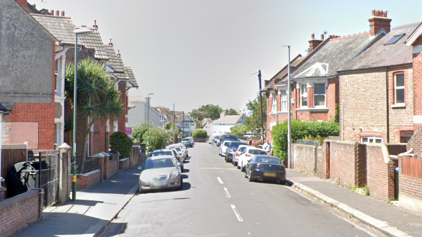 A residential street. The houses are brown brick. There are cars lining either side of the street