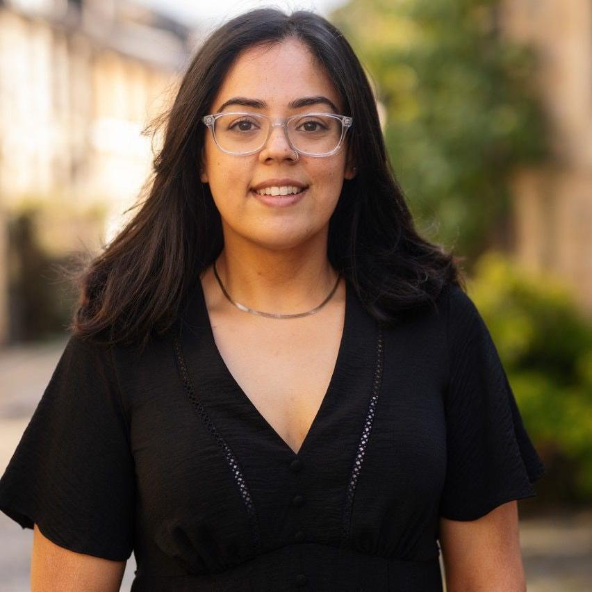 A portrait picture of Sidrah, she is a young woman and she is looking at the camera. She has black hair, black top and is wearing glasses. The trees and buildings are visible on the background.