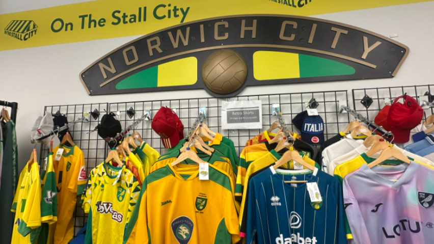 Different coloured football shirts are hung up in rows on a wall. A sign is above them which says Norwich City, and another one which has the logo for On the Stall City. 