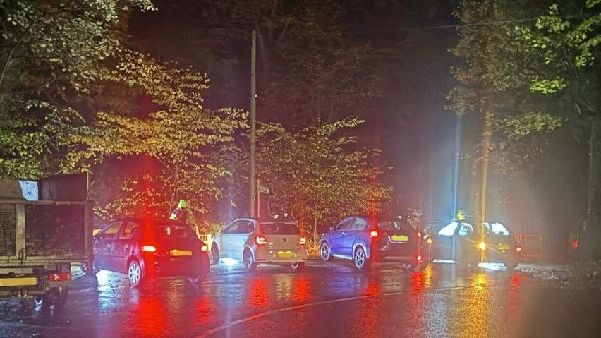 Cars lined up at the side the road on a wet evening. There lights can be seen reflecting on the wet road.