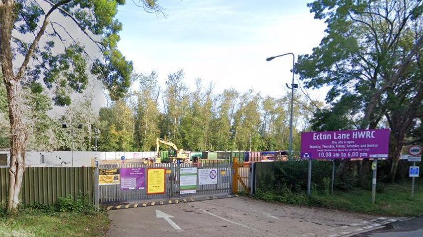 Entrance to Ecton site, showing a purple Ecton Lane HWRC sign on the right and metal gates in the centre, behind which a yellow digger is visible. There are various notices on the gates and trees behind the site.