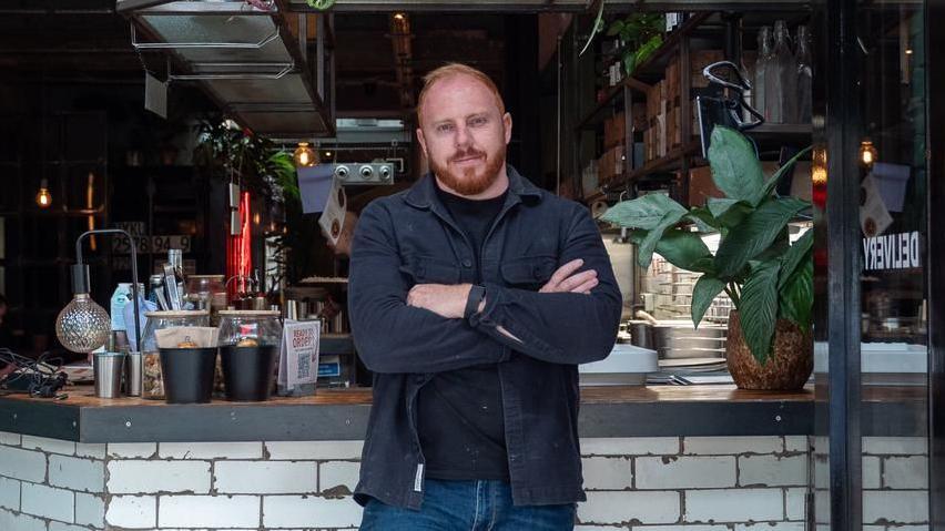 Jack Lander stands looking at the camera, arms folded, wearing an open dark shirt over a dark t-shirt, jeans, and leaned back against the food service counter at his restaurant. A plant is visible on the counter behind him, as well a lamp, and various kitchen service items.