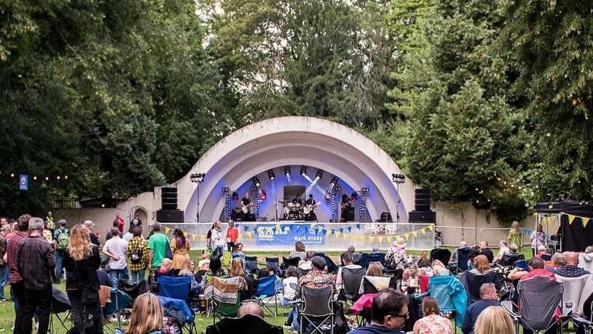 A wide shot of a semi-circle shaped stage. There is a large crowd in front of it. Many people are sat on camping chairs watching the performers on stage.