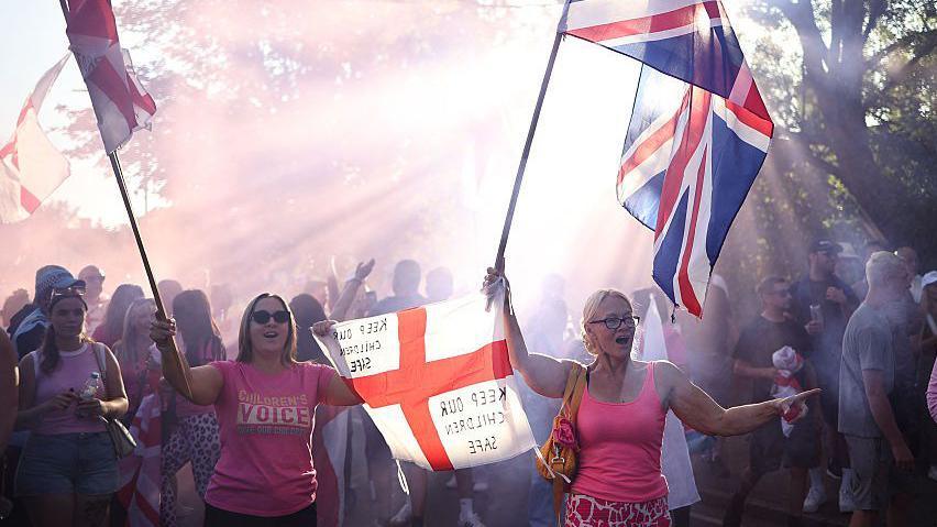 A protest outside the Bell Hotel. Two women are holding St George and Union flags. On one flag is written the message: "Keep our children safe". 