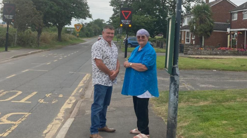 Two people looking wards camera standing in front of a Vehicle Activated Sign