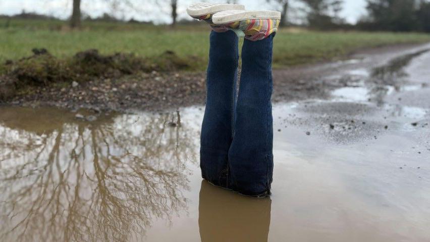 A pair of fake legs are placed in a water-filled pothole with the feet sticking out at the top. The legs are constructed from blue jeans and there are colourful shoes on the fake feet. The pothole is large and is at the side of a road. The water is muddy