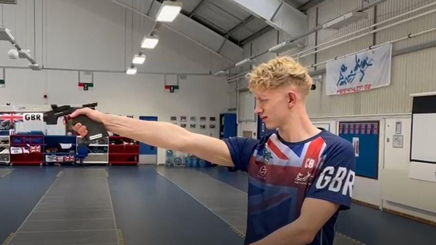 Man in blue union jack shirt holding a gun in a hall