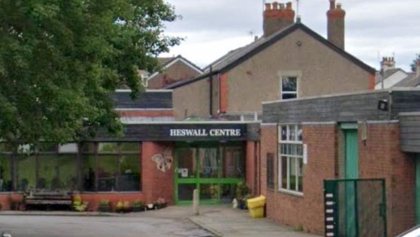 A single storey red brick building set behind trees. The sign Heswall Centre can be seen in white lettering above the green framed glass door. There is a car park with four cars parked outside the centre.