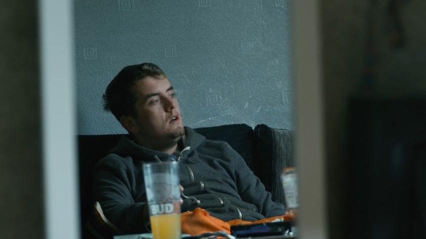 Sam is on a sofa, he is wearing a grey hoodie. He has a blank expression on his face. 