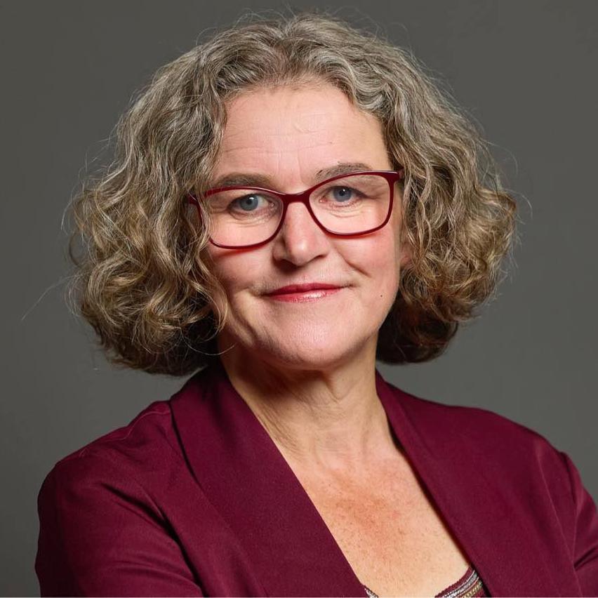 A woman stands against a grey background. She has curly grey hair and glasses. She is wearing a maroon jacket.