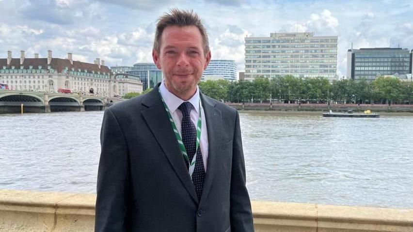 David Williams, wearing a suit, stands by a river. Buildings can be seen on the other side of the water.