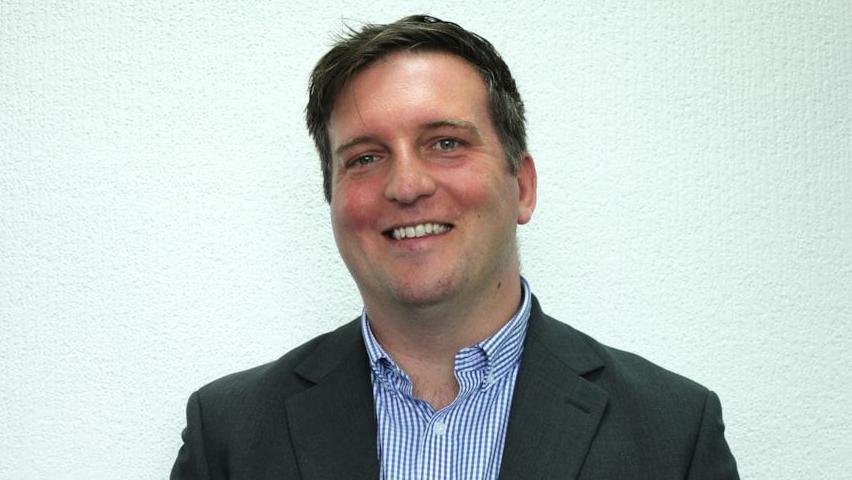 A head and shoulders picture of James Hanson, shown standing in front of a light green wall and smiling at the camera. He has short dark hair and is wearing a grey jacket and white and blue checked shirt. 