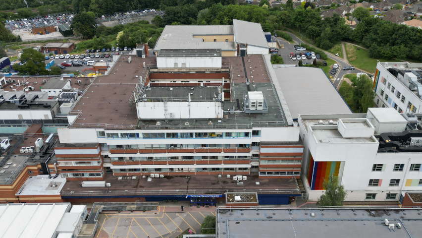 A drone picture has been used to show what the hospital looks like from above. The building is square shaped, and it is four to five storeys high. It is mainly white, with red accents. 