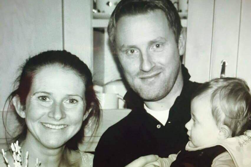 A black and white image of Rhian with her husband Paul and baby George. Rhian and Paul are looking at the camera and George is looking at Rhian. Rhian has her dark hair pulled back, Paul has short fair hair.