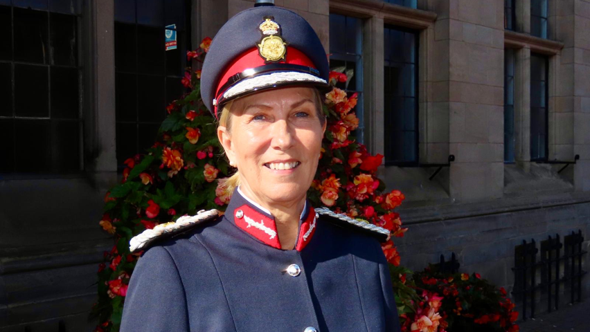 A woman stands and smiles in front of a building and a large bush with red flowers. She is wearing a dark blue lord-lieutenant's hat and uniform.