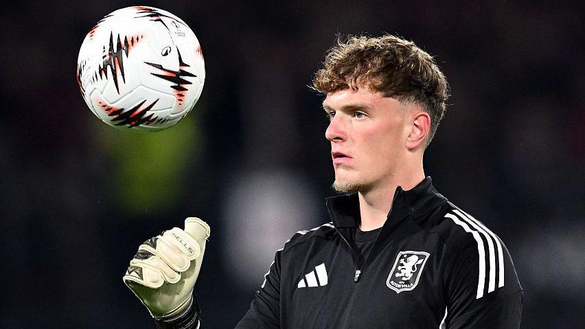 Sam Proctor throws a ball during a warm-up session for Aston Villa. He is wearing a black club zip top.