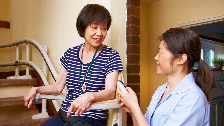 Woman sitting on stairlift at bottom of a flight of stairs, being helped by a female nurse